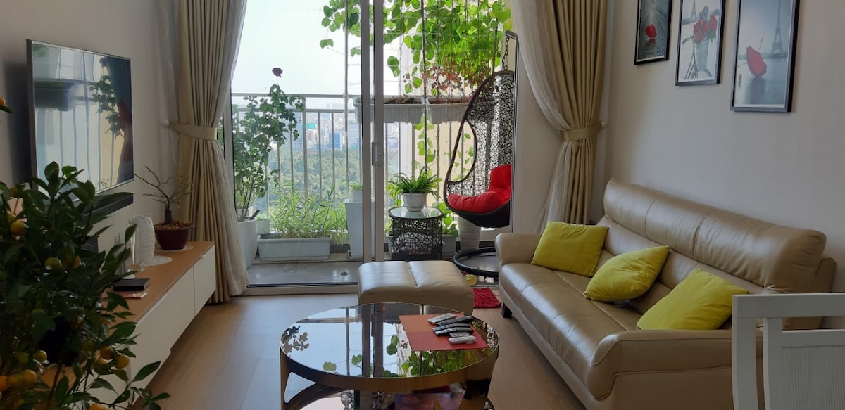 A bright living area features a comfortable tan sofa adorned with yellow cushions. A glass coffee table sits in front, while a hanging chair adds a unique touch. Floor-to-ceiling windows frame lush greenery outside, enhancing the indoor space's openness and natural light.