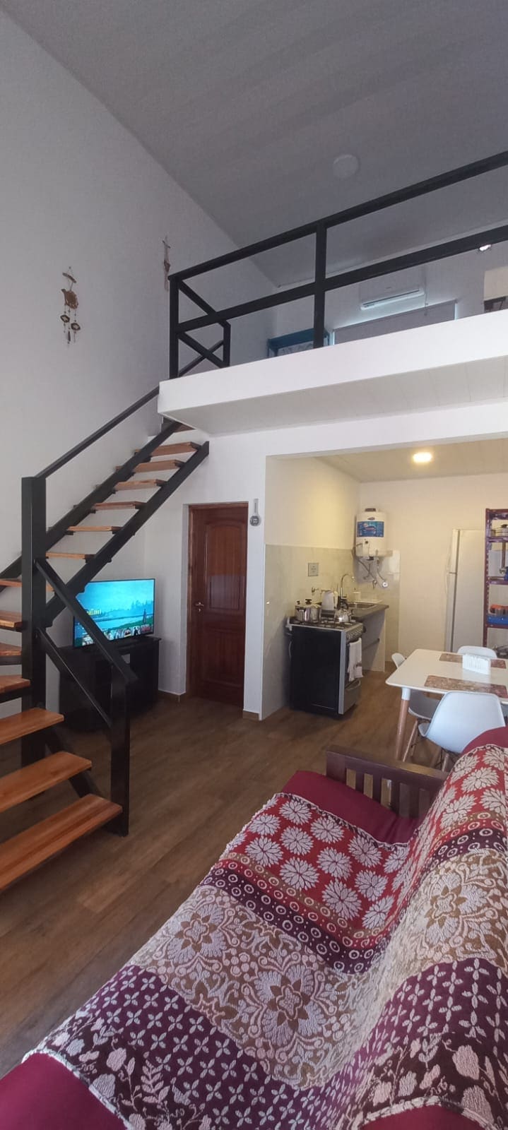 A modern living space is shown, featuring a staircase leading to an upper level. A cozy seating area is present with a patterned sofa, while a TV sits on a cabinet. The kitchen area is visible, equipped with essential appliances, and a dining area is set nearby.
