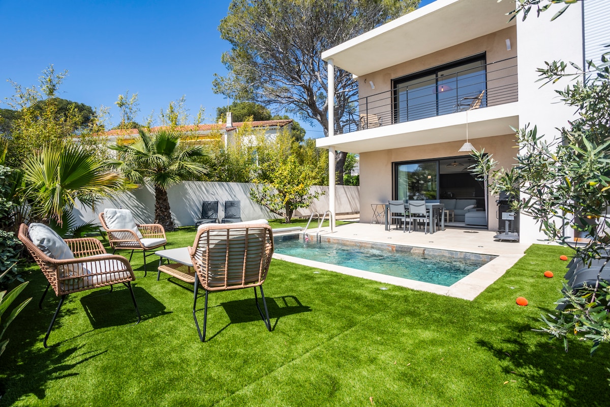 An outdoor area features a private stone pool surrounded by well-kept artificial grass. Comfortable seating is provided by a set of chairs and a sofa. A dining table is positioned near the house, with lush greenery and trees creating a serene atmosphere.