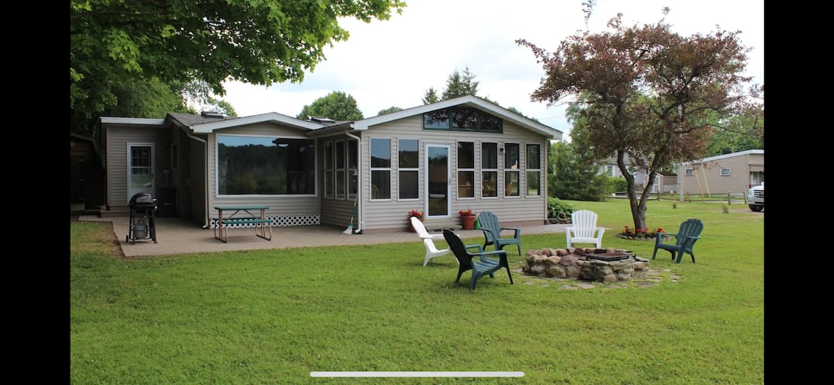 A spacious lakefront cottage is surrounded by lush green lawn. A cozy fire pit area features several chairs, while large windows allow for natural light. A picnic table is positioned nearby, enhancing the outdoor space for relaxation and gatherings.