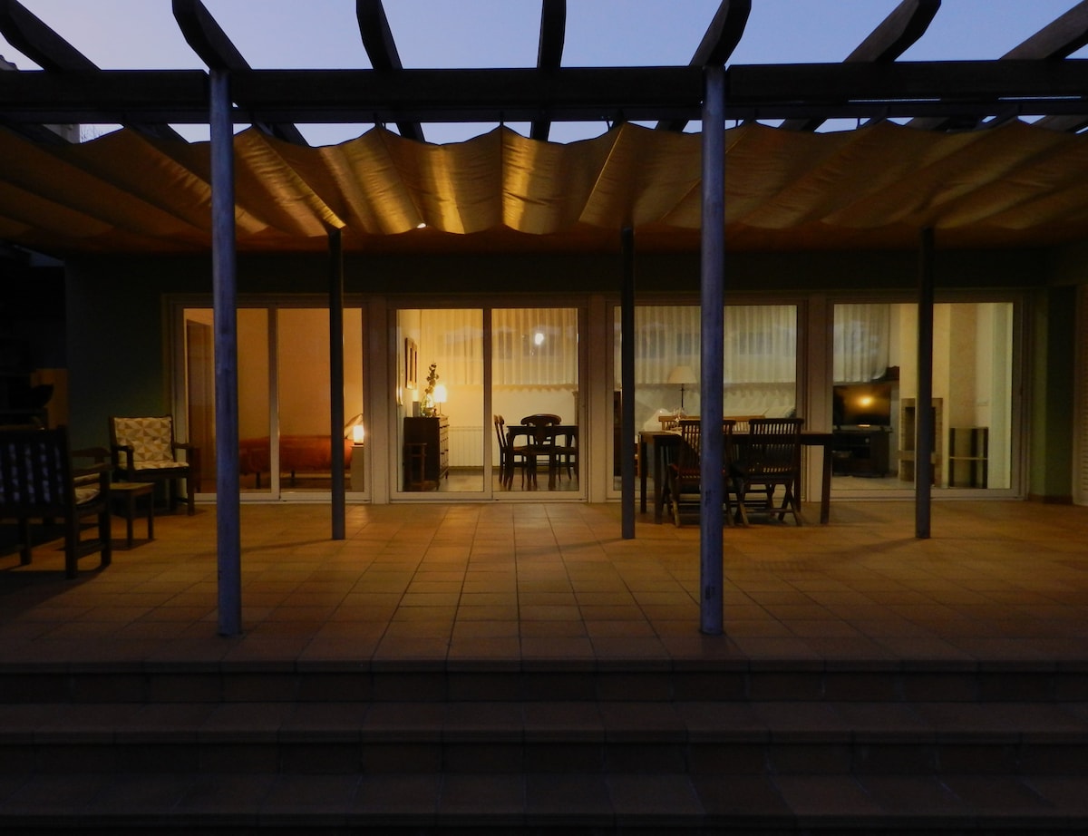 An outdoor terrace is visible, featuring a partially covered seating area with wooden furniture. The large glass doors offer a view into a well-lit living space, where warm lighting highlights the inviting interiors. Soft shadows are cast as dusk approaches.