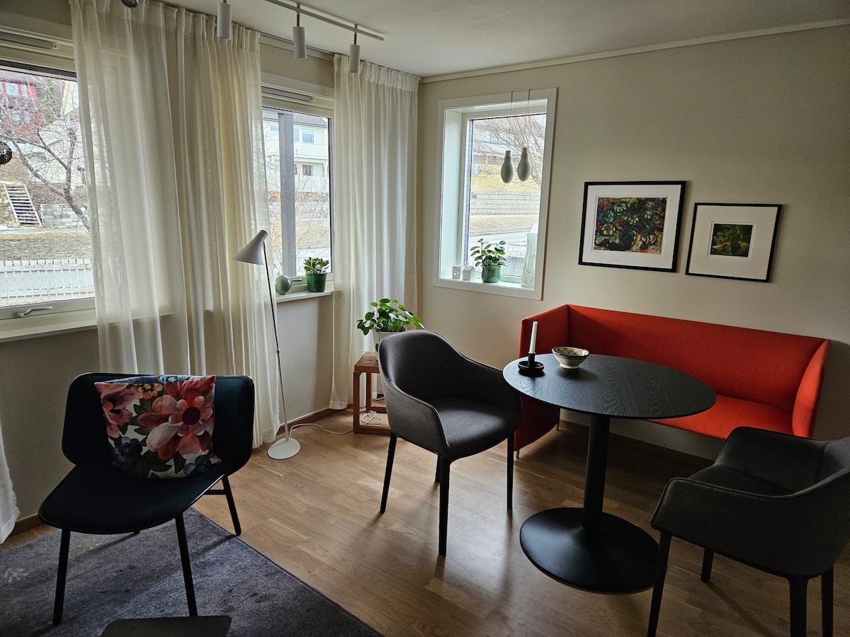 The living area is brightened by natural light streaming through two windows, complemented by sheer curtains. A round table sits in the center, surrounded by various chairs, including an orange bench. Two framed artworks add visual interest to the neutral-toned walls.