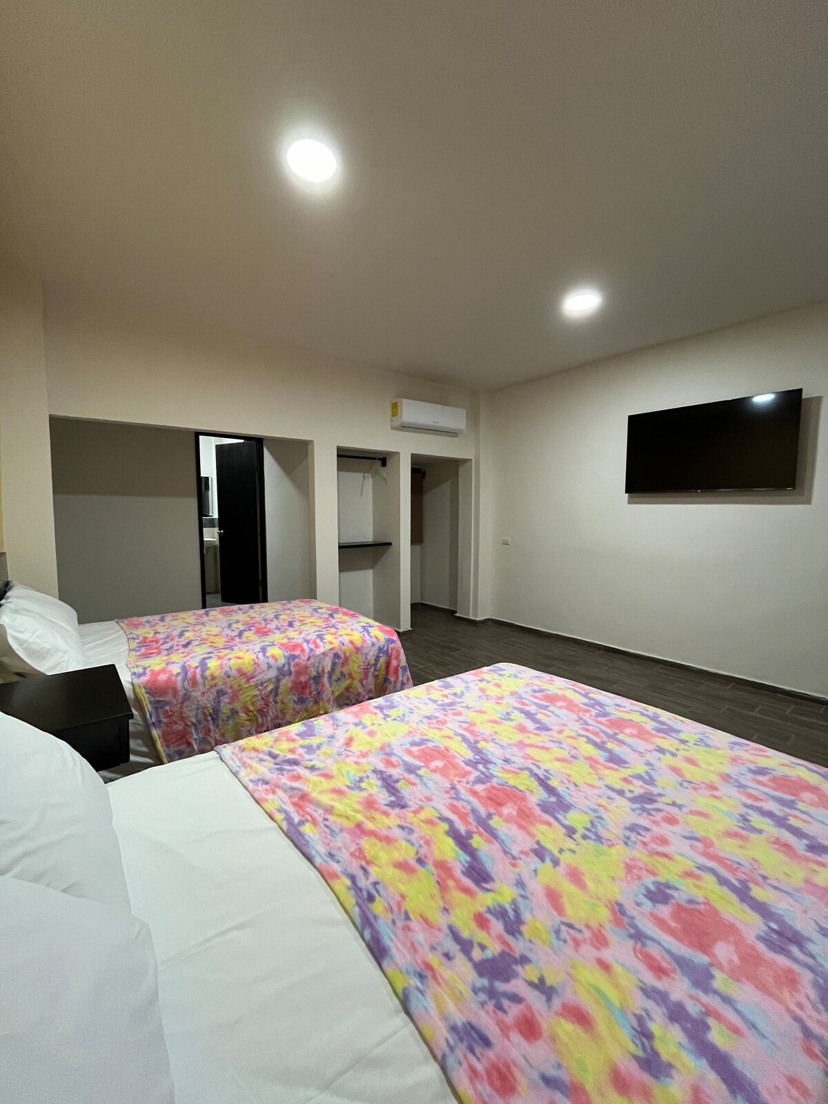 A spacious room is presented with two beds, each covered in a colorful floral-patterned blanket. A mounted television is visible on the wall, and a closet area is located in the background, providing ample storage space. Soft lighting illuminates the warm-toned walls.