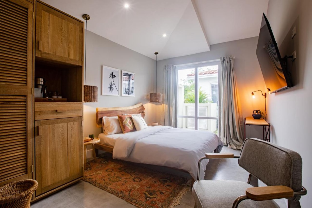 A well-appointed studio unit features a spacious bed with soft linens, complemented by wooden furnishings. A cozy chair and a decorative area rug add warmth. Natural light enters through large windows, while artistic prints adorn the walls, enhancing the inviting ambiance.