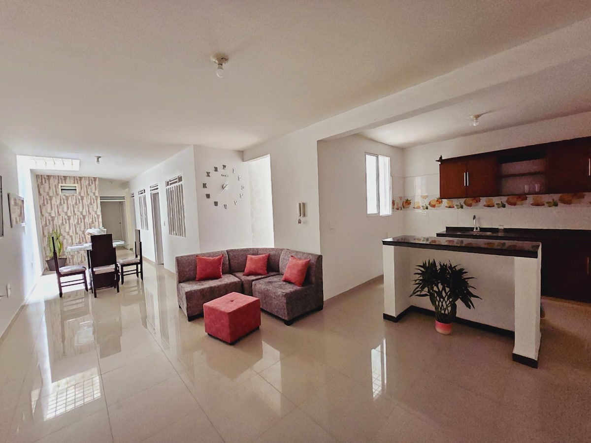 A spacious living area features a comfortable gray sofa with pink accent cushions and a matching ottoman. A dining table is positioned in the background. The kitchen showcases dark cabinetry, with ample natural light provided by multiple windows.