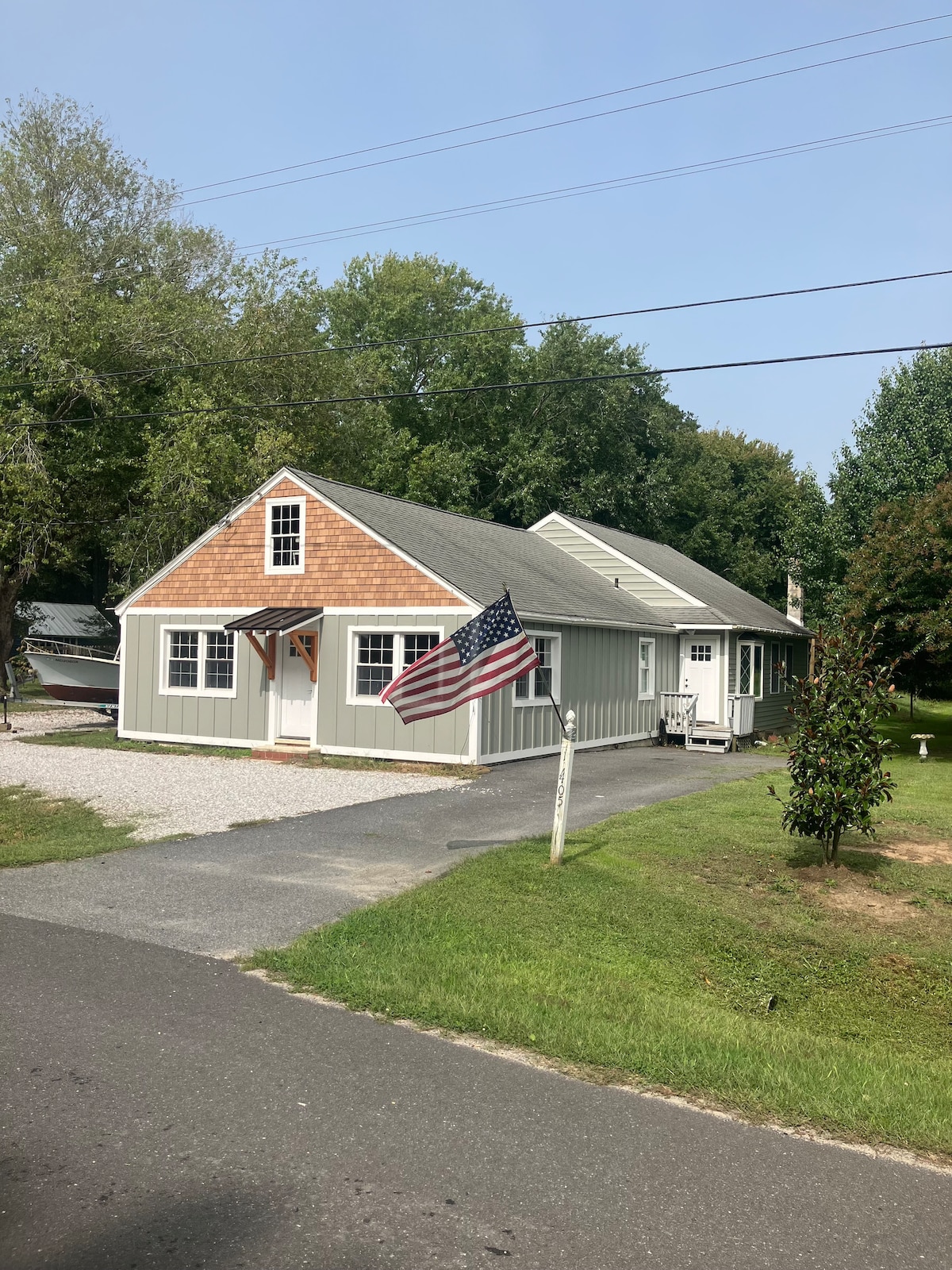 The cottage features a charming exterior with a combination of wood and siding. A gabled roof, traditional shingles, and a welcoming entrance are visible. An American flag is displayed prominently by the entrance, and the surrounding greenery enhances the peaceful setting.
