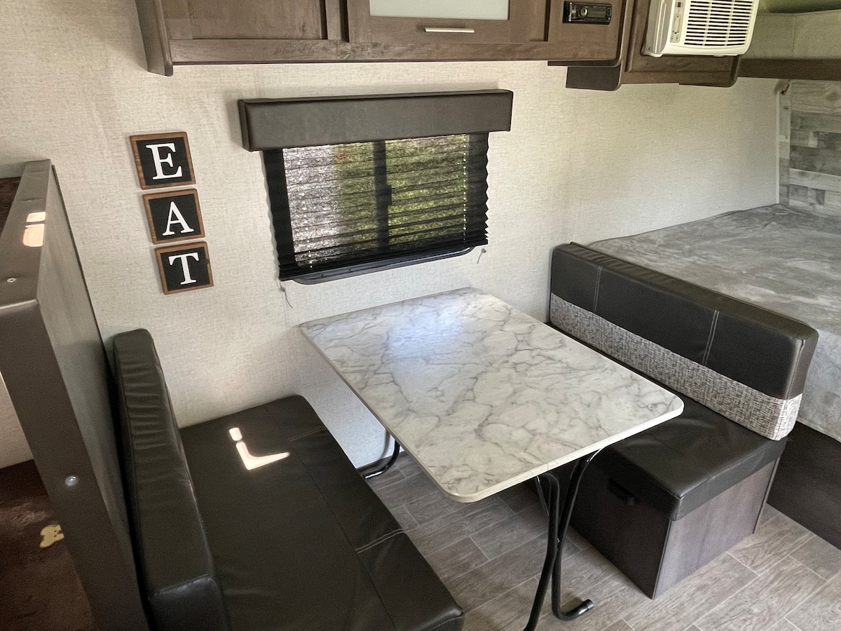 A cozy dinette area is featured with a marble-top table surrounded by dark upholstered seating. A window with closed black blinds allows natural light to filter in, while the wall displays wooden letters spelling 'EAT.' The nearby sleeping area is visible in the background.