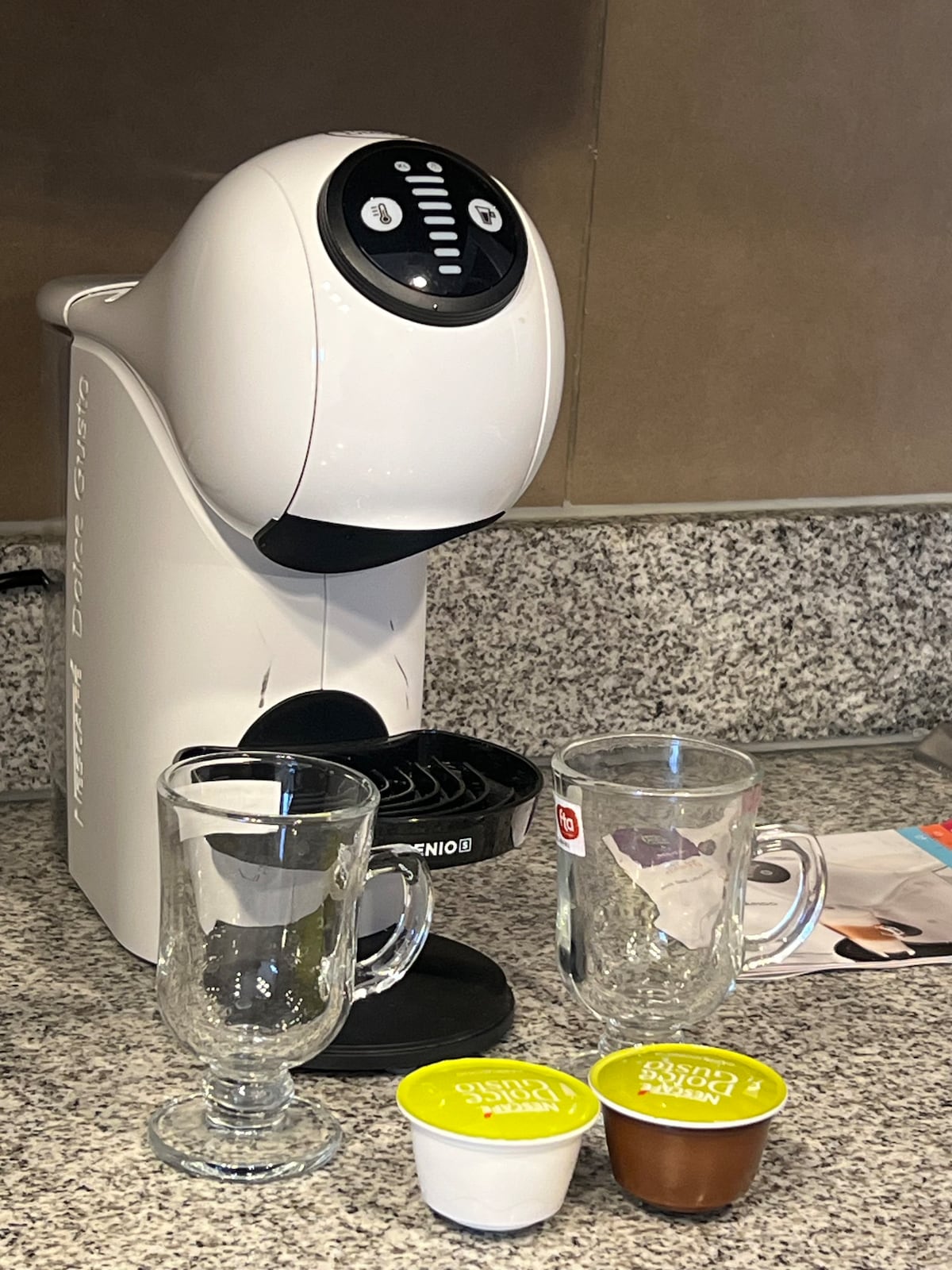 A coffee maker is positioned on a speckled granite countertop. Two clear glass mugs are placed next to the machine, alongside coffee pods in white and green packaging. An informational brochure is partially visible in the background.