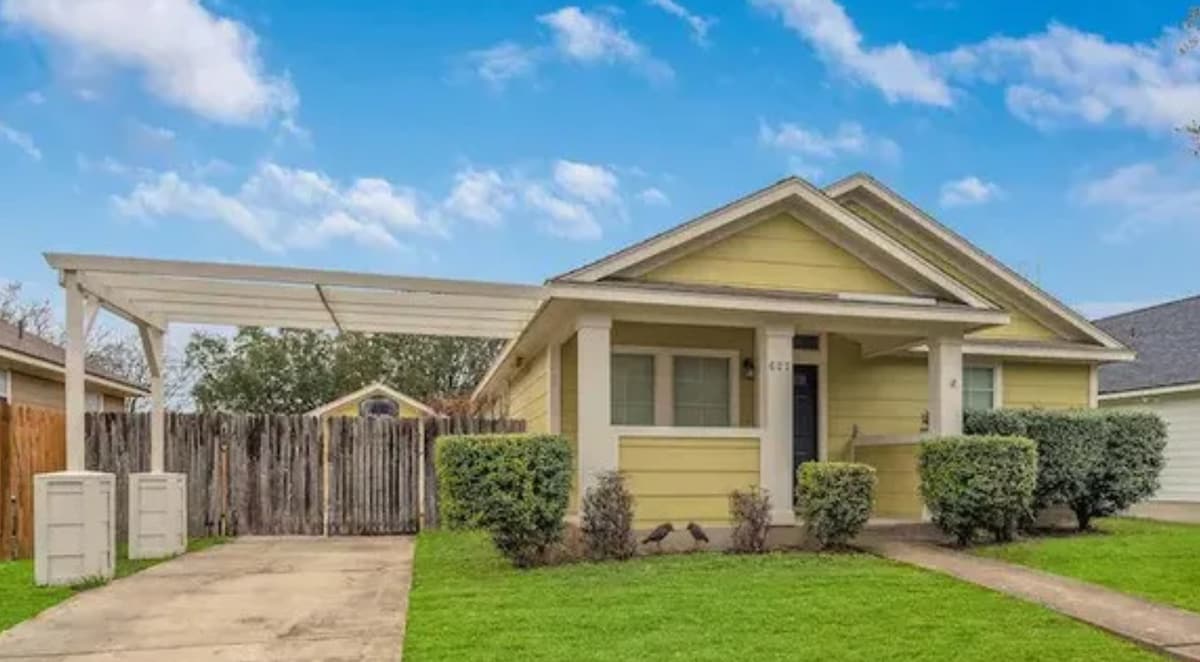 A one-story home with a light yellow exterior is showcased, featuring a carport and a clear pathway leading to the entrance. Neatly trimmed bushes line the walkway, and a wooden fence is visible in the background, providing privacy. The lawn is green and well-maintained.