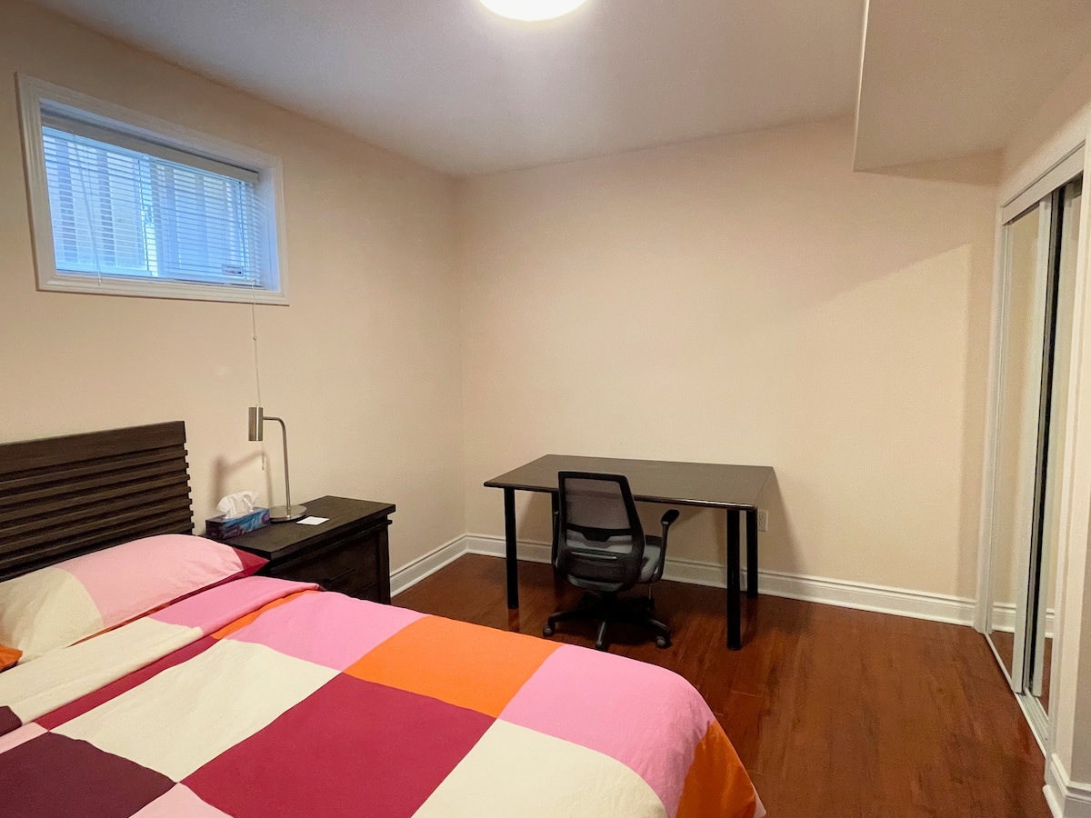 A private room features a queen size bed with a multicolored quilt, complemented by a bedside lamp and a wooden side table. A simple desk with an office chair is positioned against a wall. Natural light enters through a small window, and mirrored sliding doors are visible.