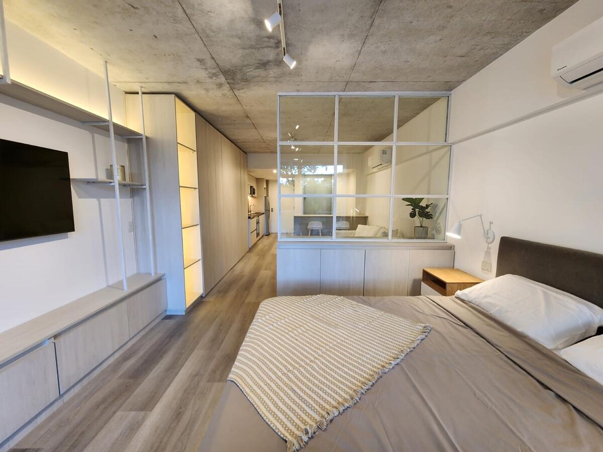 An open layout is seen featuring a cozy sleeping area with a spacious bed and bedside lamp. A glass partition separates the living space from the kitchen, which is partially visible in the background. Natural light illuminates the room, creating a bright atmosphere.