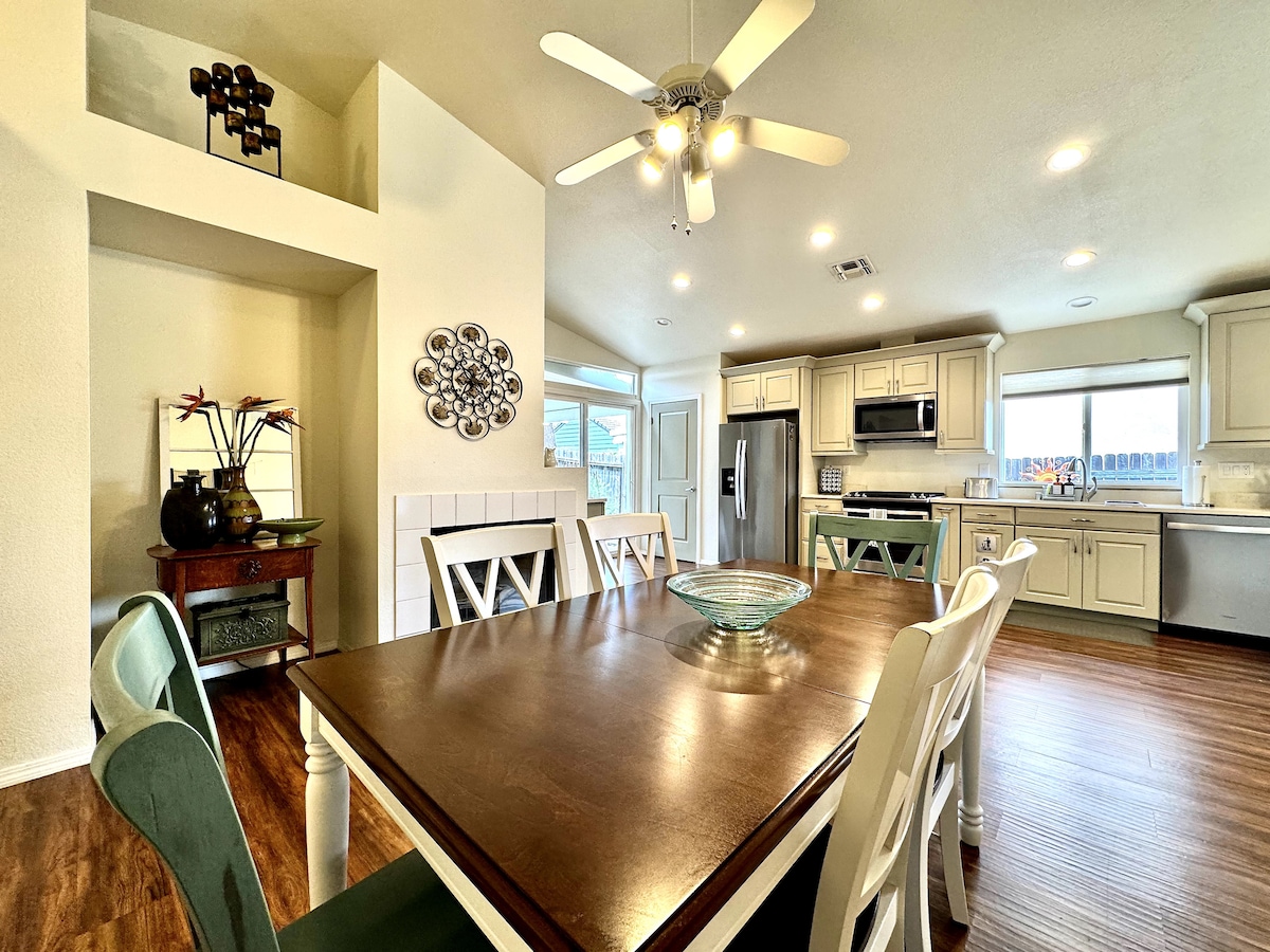 A spacious dining area is highlighted by a large wooden table surrounded by six chairs. The kitchen is visible in the background, featuring modern appliances and a large window that provides natural light. A ceiling fan adds functionality, while decorative elements are placed on the wall.
