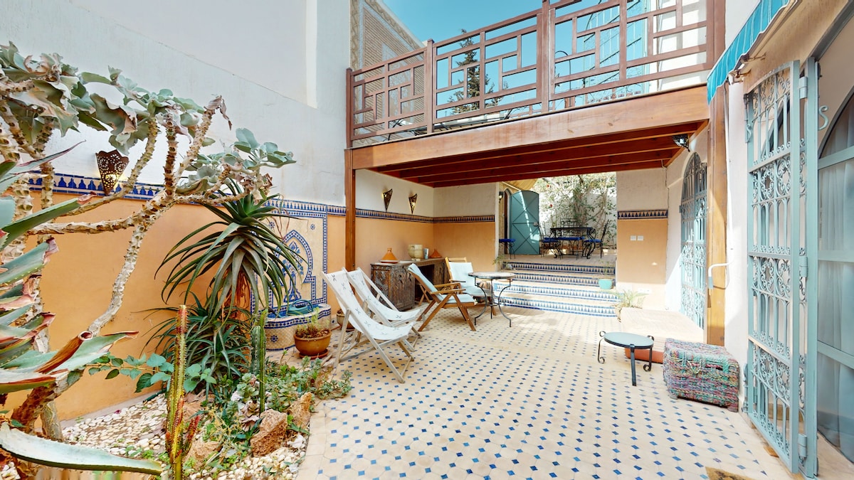 An outdoor terrace is surrounded by decorative tiles and lush greenery. A seating area is highlighted by comfortable loungers and a wooden table. A small pool is visible in the background, complemented by a staircase leading to an upper level.
