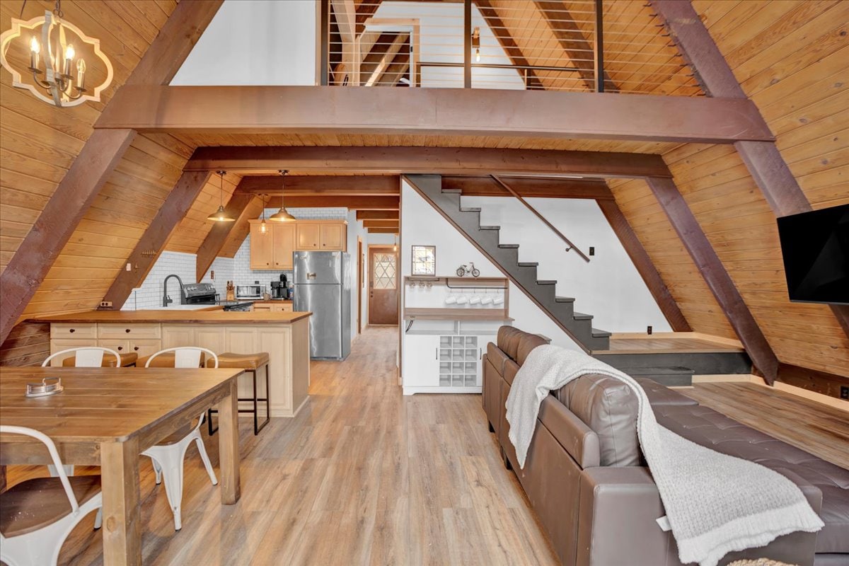 An open-concept living space features vaulted wood ceilings and a mix of rustic and modern design elements. A dining table is positioned near the kitchen, which includes a stainless steel refrigerator. Stairs lead to a loft area, while a comfortable couch provides a cozy seating option.