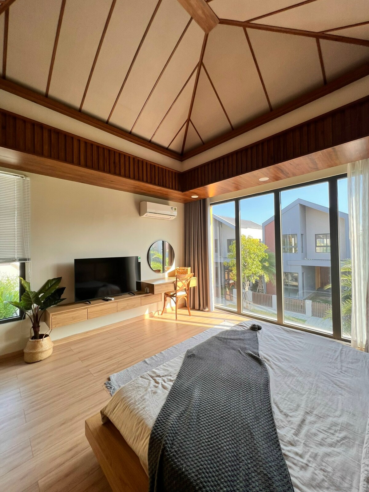 A spacious bedroom features a large window that allows natural light to fill the room. A comfortable bed with a light blanket is paired with a small armchair and a TV mounted on a wooden cabinet. Green indoor plants add a touch of nature.