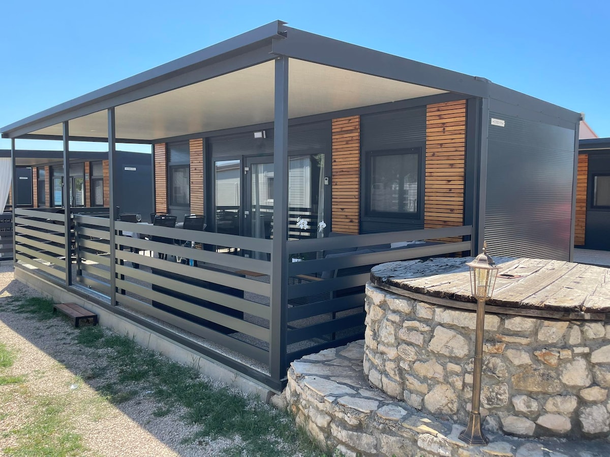 The exterior of the mobile home is showcased, featuring a large, covered terrace with contemporary railings. A stone wall surrounding a circular feature is visible in the foreground, complementing the structure's modern design. Clear blue skies enhance the inviting environment.