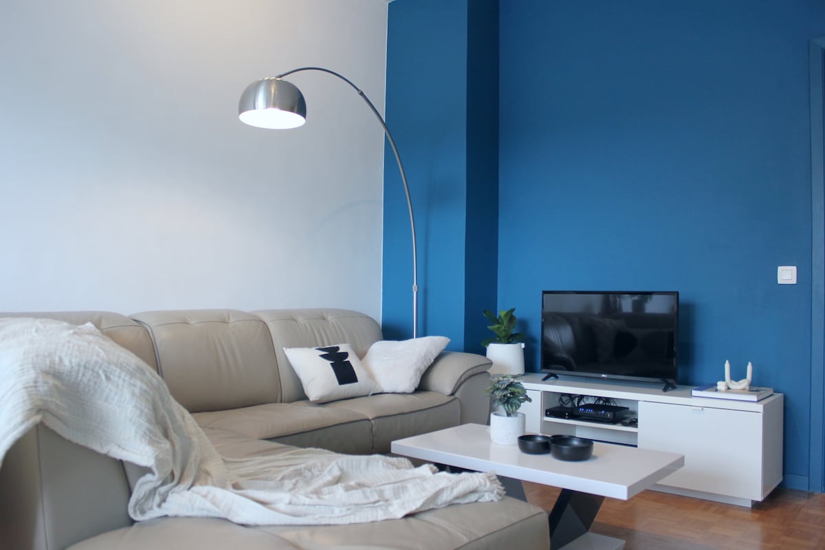 A cozy living area is presented with a light-colored sectional sofa, a modern floor lamp, and a television on a minimalist white console. A small coffee table with decorative bowls is positioned in front, while a green plant adds a touch of freshness to the space.