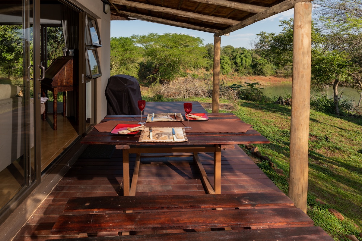 An outdoor dining area features a wooden table set with plates and glasses, overlooking a lush landscape. The space is sheltered by a wooden roof, with glass doors visible in the background leading to the interior. Natural light illuminates the serene surroundings.