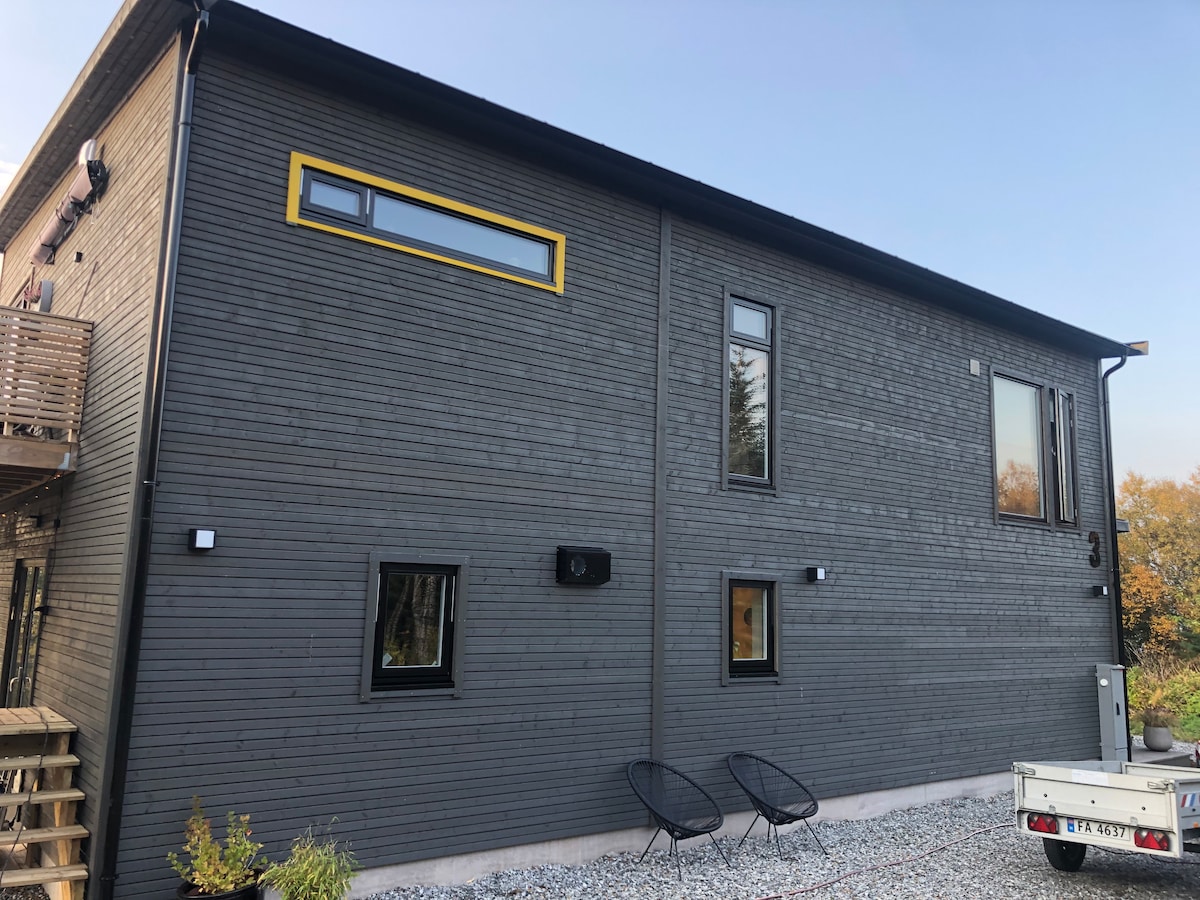 The exterior of the house features a modern design with a dark wooden facade. Large windows provide natural light, while two round chairs are placed outside on gravel. A small trailer is visible nearby, complementing the natural surroundings.