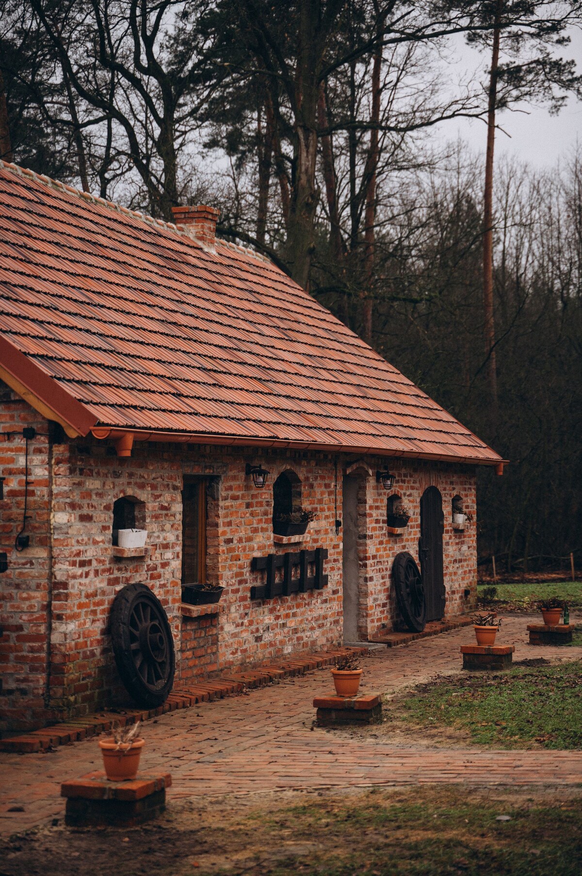 A charming brick cottage is framed by a natural woodland setting, featuring a warm red roof and rustic wooden accents. Potted plants are arranged along the walkway, which is made from stone, leading up to the entrance that showcases traditional wagon wheels as decor.