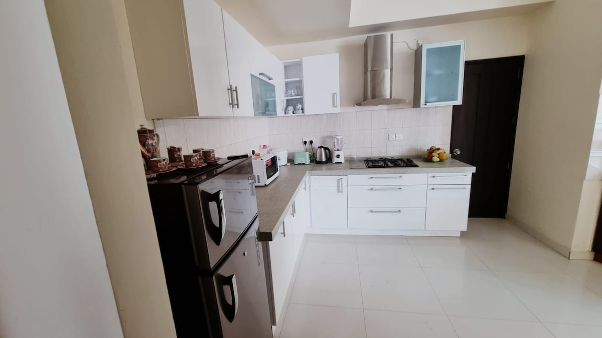 A modern kitchen features sleek white cabinetry and a spacious countertop. Appliances include a refrigerator, a coffee maker, and a stove, all designed for convenience. Natural light brightens the space, enhancing its functional layout, which accommodates meal preparation for families.