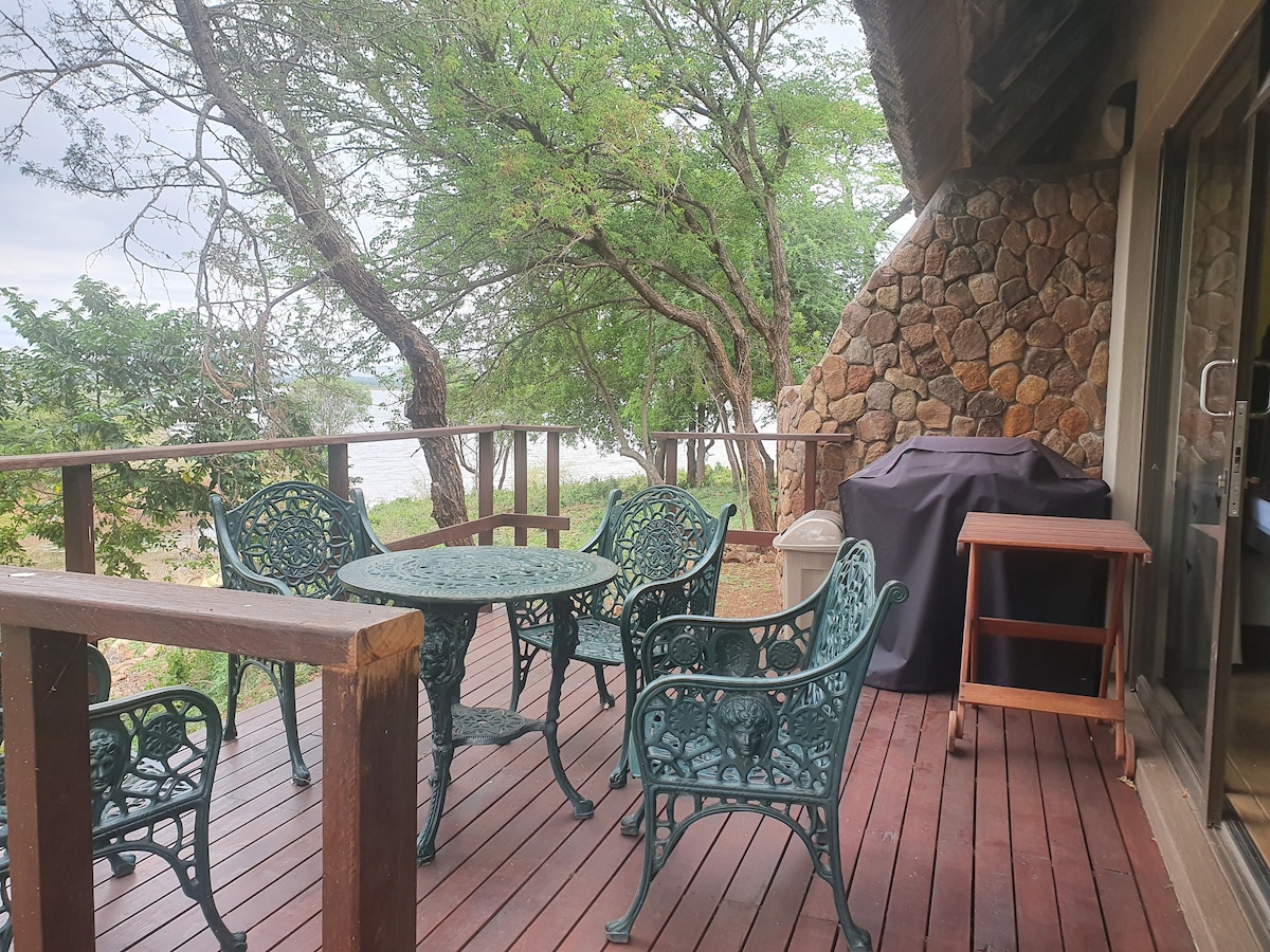 A spacious wooden deck features a round table surrounded by four intricately designed chairs. A gas braai is positioned on one side, with trees providing a natural backdrop and views of the nearby inlet in the distance.