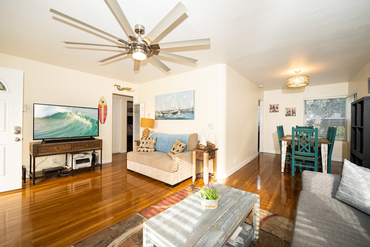 A spacious living area is featured with polished wooden floors and a ceiling fan. A large flat-screen television is positioned near a comfortable beige sofa. A dining table with teal chairs is also present, complemented by a decorative area rug and natural light from a nearby window.