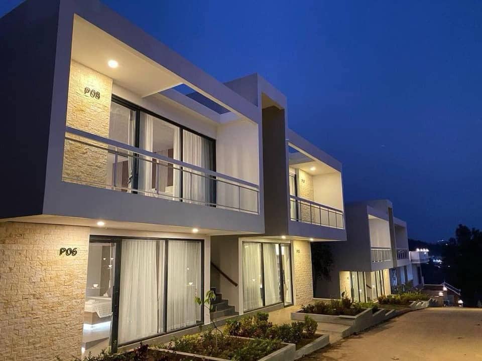 Modern two-story villas are situated under a twilight sky, featuring balcony access topped with sleek railings. Large glass doors illuminate the interiors, while neatly trimmed landscaping enhances the aesthetic near the entrance.