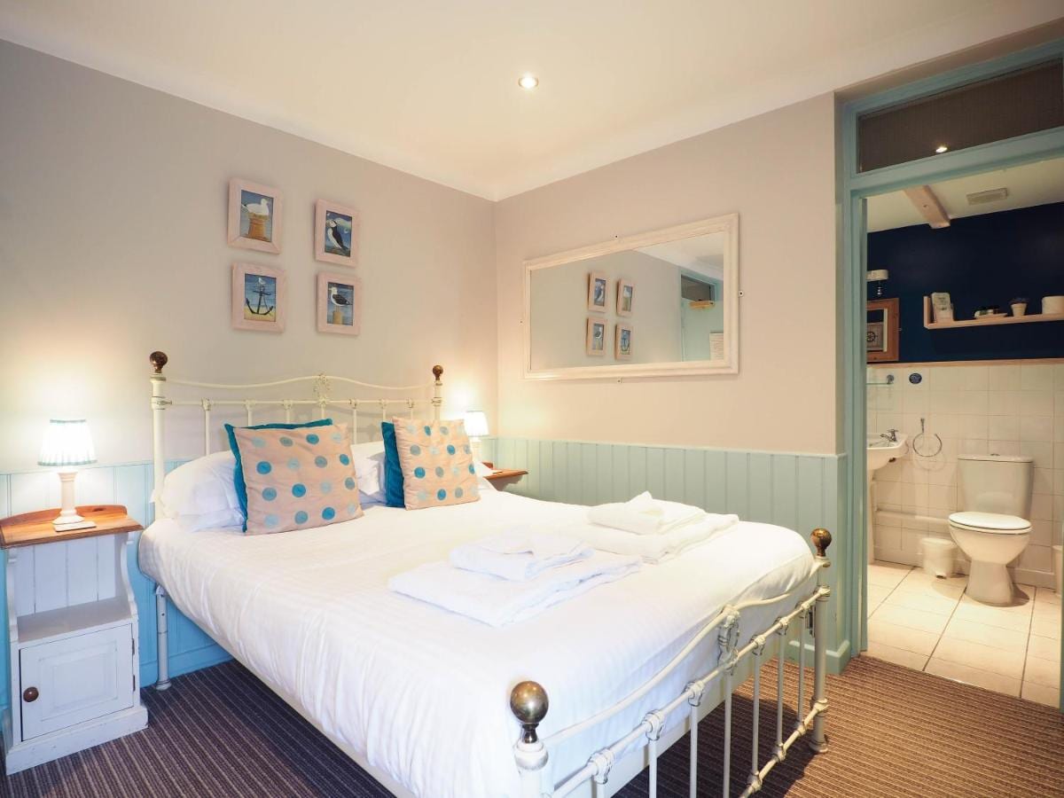 A well-lit bedroom features a spacious bed dressed in white linens, adorned with decorative pillows. Two bedside lamps provide soft lighting. Framed pictures are displayed on the wall, and a mirror reflects the calm space. An ensuite bathroom is visible in the background.