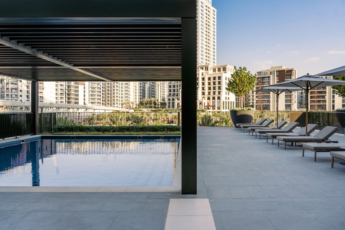A tranquil swimming pool is framed by a modern pavilion with a sleek roof. Lounge chairs are arranged around the pool on a spacious tiled deck, surrounded by greenery and city views. Clear skies provide a bright backdrop to the serene setting.