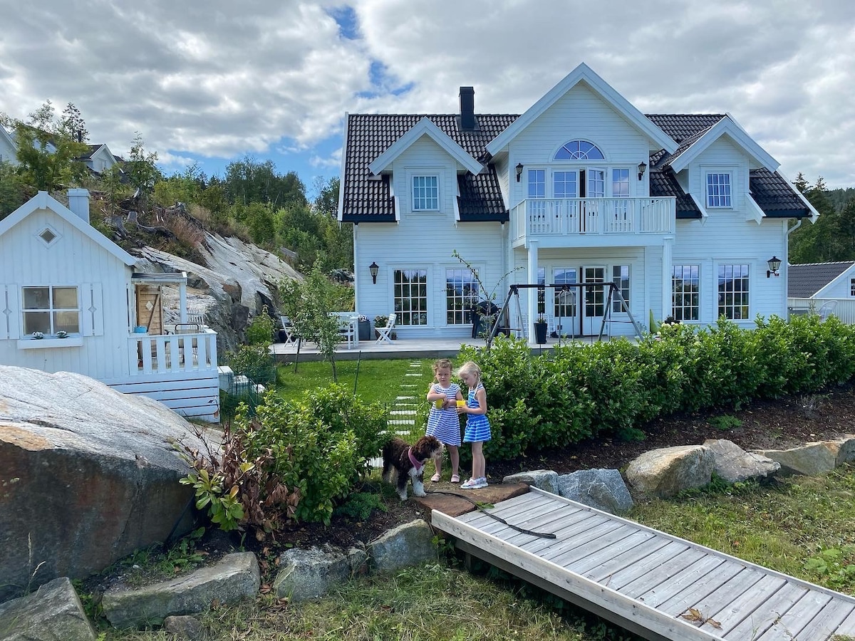 A charming white house with large windows is set against a cloudy sky. Two children are seen playing in the foreground, surrounded by green shrubbery and a wooden walkway. A smaller playhouse stands nearby, contributing to the idyllic outdoor environment.