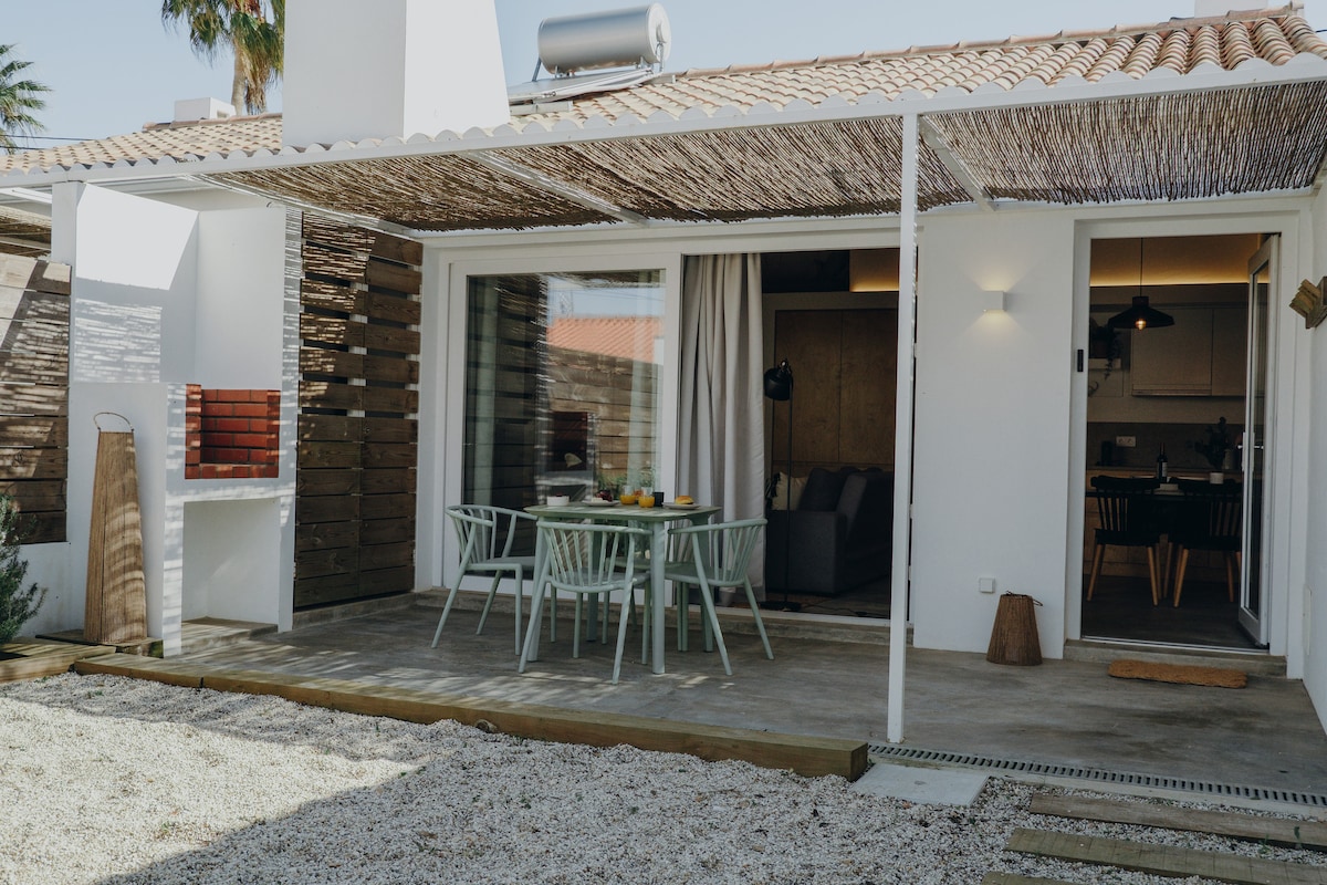 An outdoor patio area features a dining table surrounded by light green chairs, offering a space for meals and relaxation. A barbecue area is visible, with a cozy seating arrangement just inside the glass doors leading to the interior living space.