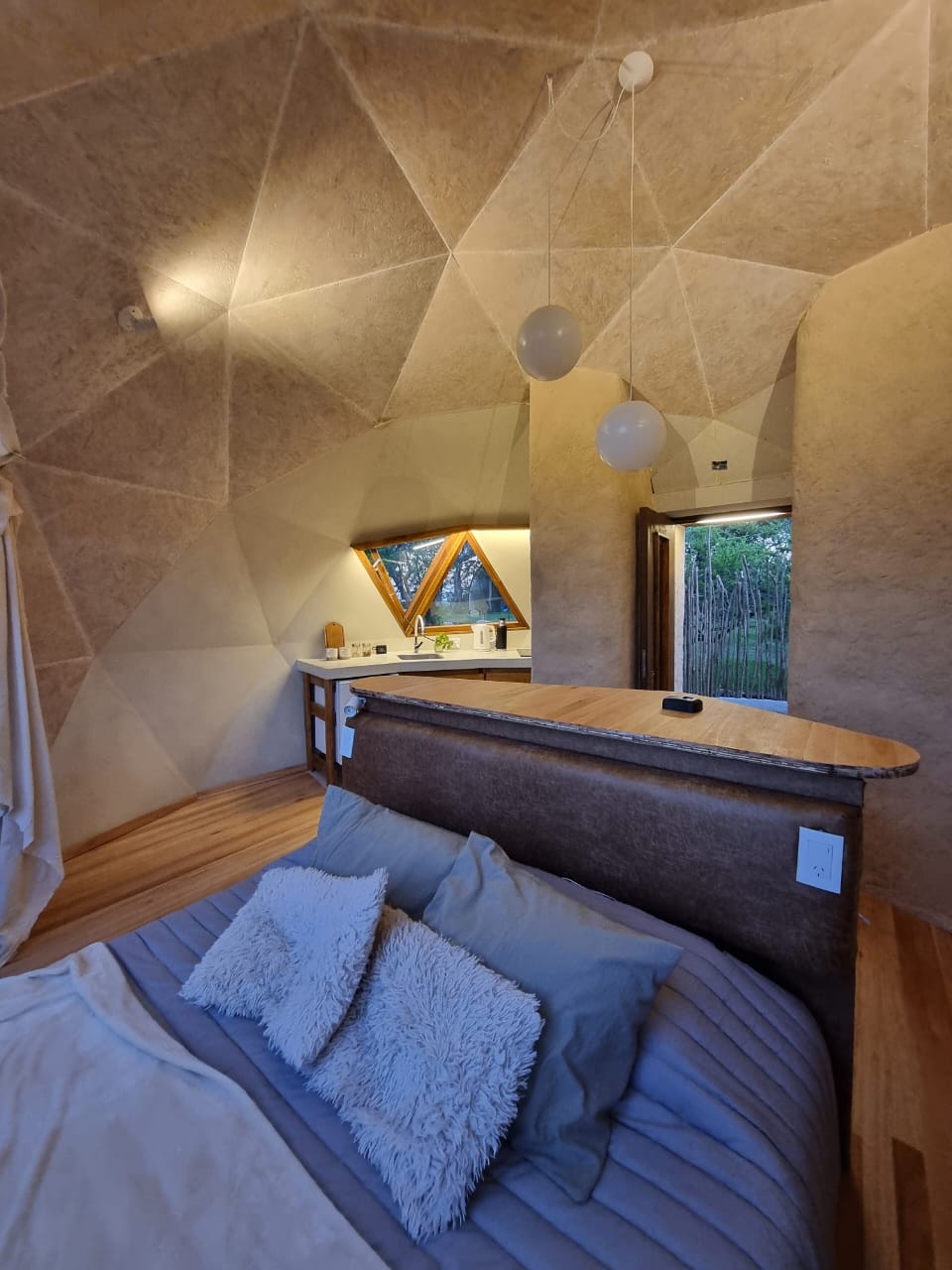A uniquely shaped interior showcases a cozy bed with plush pillows, draped in soft bedding. A kitchen area is visible in the background, featuring wooden surfaces and triangular windows that invite natural light, blending comfort with a modern design.