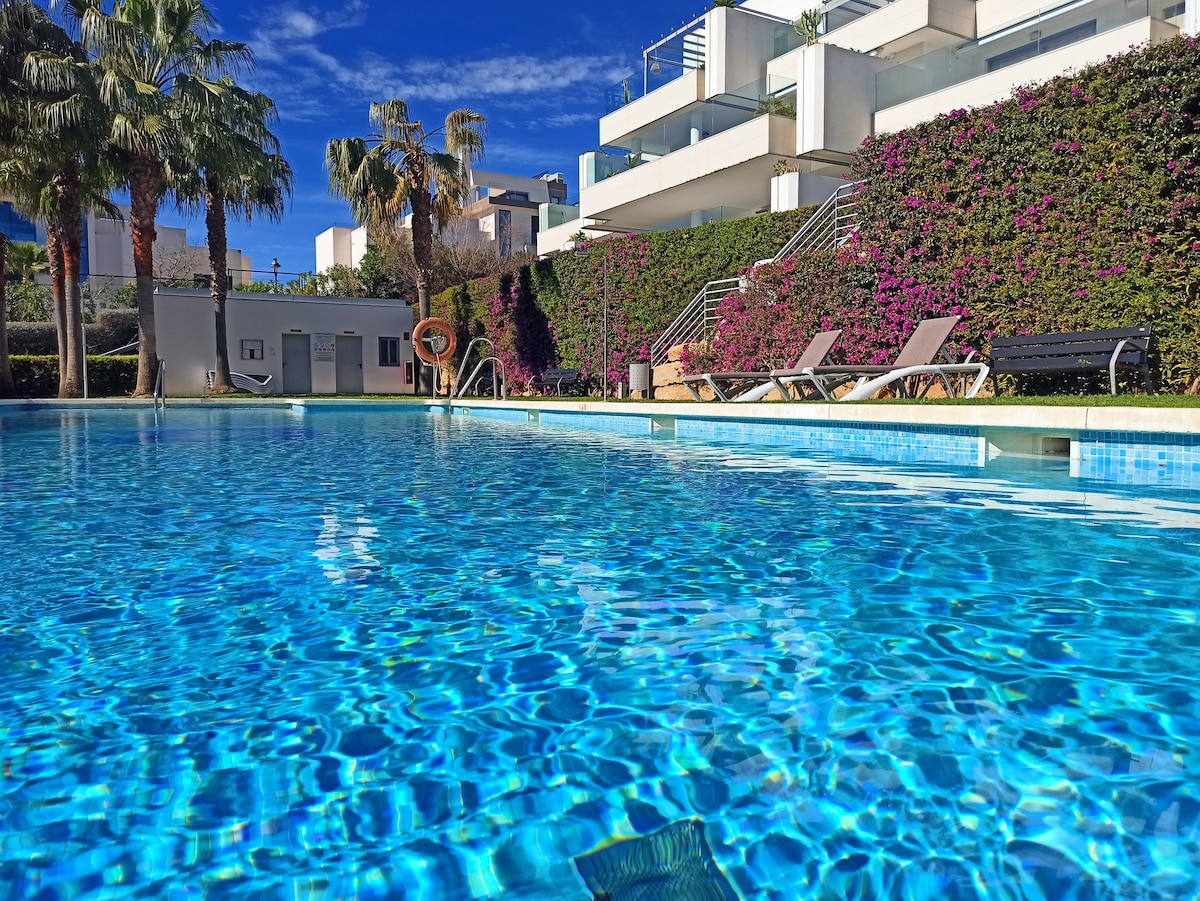 A sparkling shared pool reflects the bright sky and surrounding greenery. Sun loungers are positioned along the pool's edge, while vibrant plants add a touch of color to the tranquil environment.