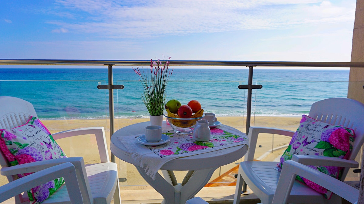 A small balcony table is set with a fruit bowl and coffee cups, surrounded by two white chairs. The ocean view stretches beyond, with gentle waves lapping at the sandy shore. Soft, pastel cushions add a touch of comfort to the seating area.