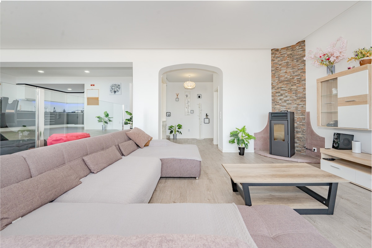 The spacious living area features a modern sectional sofa, a wooden coffee table, and a stylish stone accent wall. Potted plants add a touch of greenery, while large windows allow for ample natural light. A cozy fireplace is positioned nearby, contributing to the room's warmth.