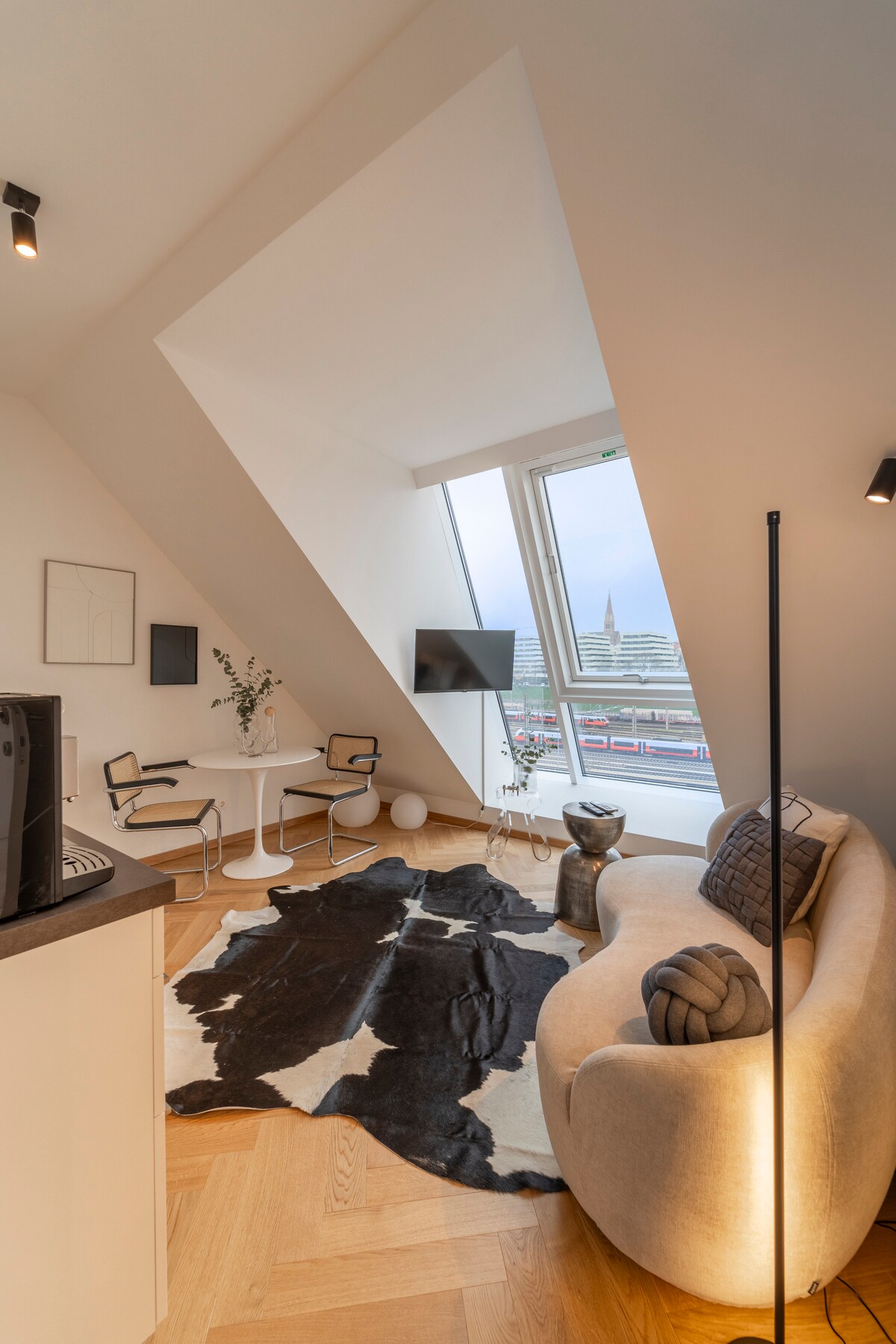 A modern living space features a uniquely angled window that offers a city view. A light-colored sofa is complemented by a decorative throw and pillow. A small, round dining table with two chairs occupies one corner, while a sleek TV is mounted on the wall.