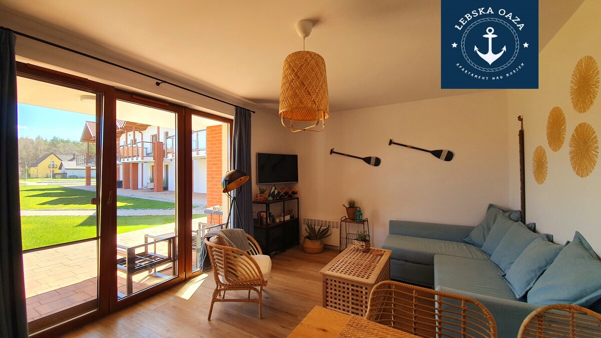 A bright living area features a light blue sectional sofa and a rattan coffee table. Large sliding doors open to a patio overlooking a grassy area. Two paddles are mounted on the wall, and natural light fills the space from multiple windows.