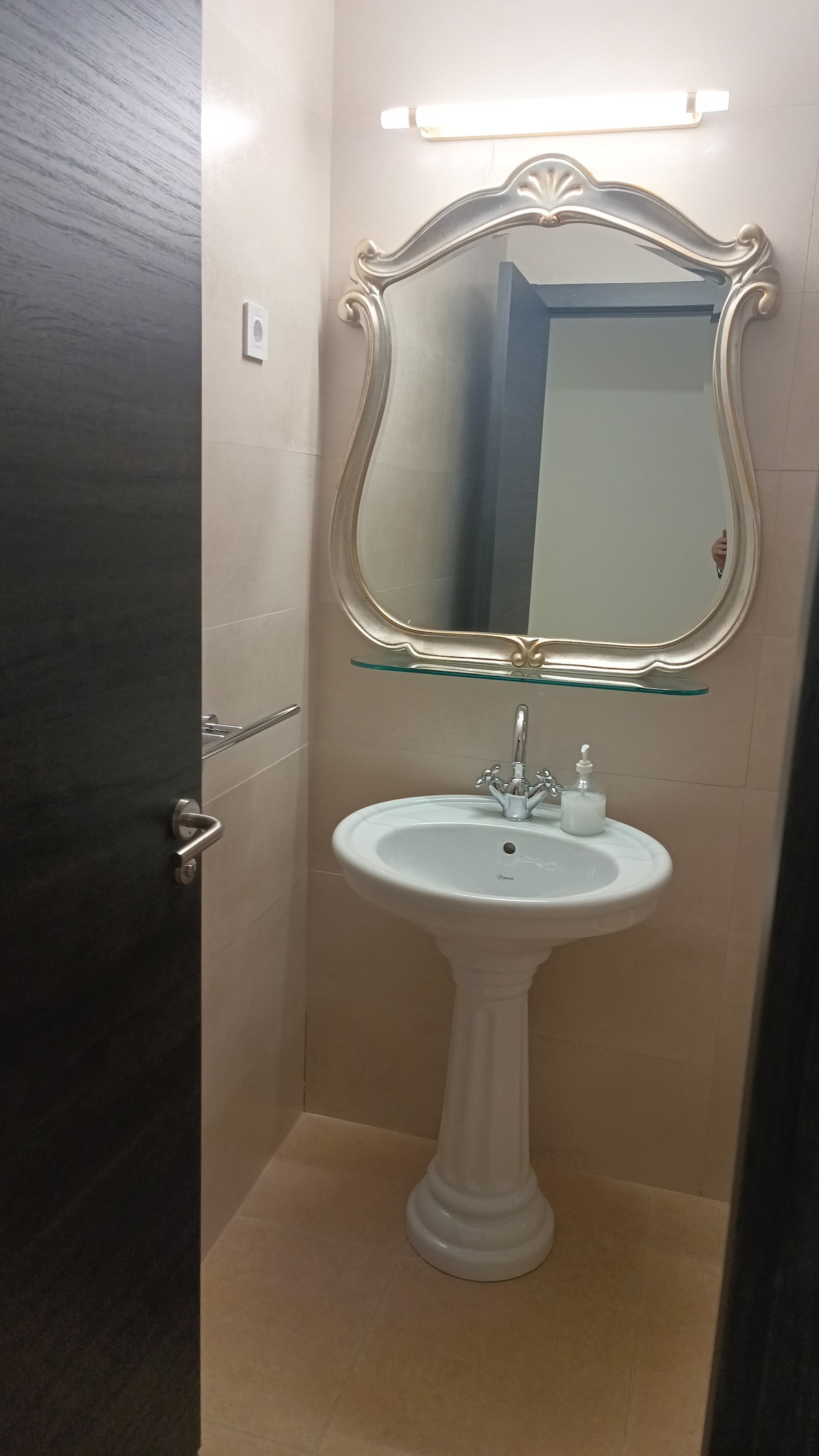 A small bathroom is showcased, featuring a white pedestal sink with a vintage-style mirror above it. Soft lighting illuminates the space, while beige tiles line the walls and floor. A hand towel rack is visible next to the sink.