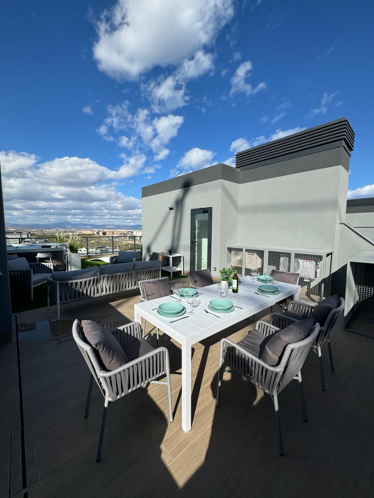 A rooftop terrace is showcased, featuring a dining table set for six with light green plates and glassware. Comfortable grey chairs surround the table. The background reveals a clear blue sky with scattered clouds and scenic views of the surrounding landscape.