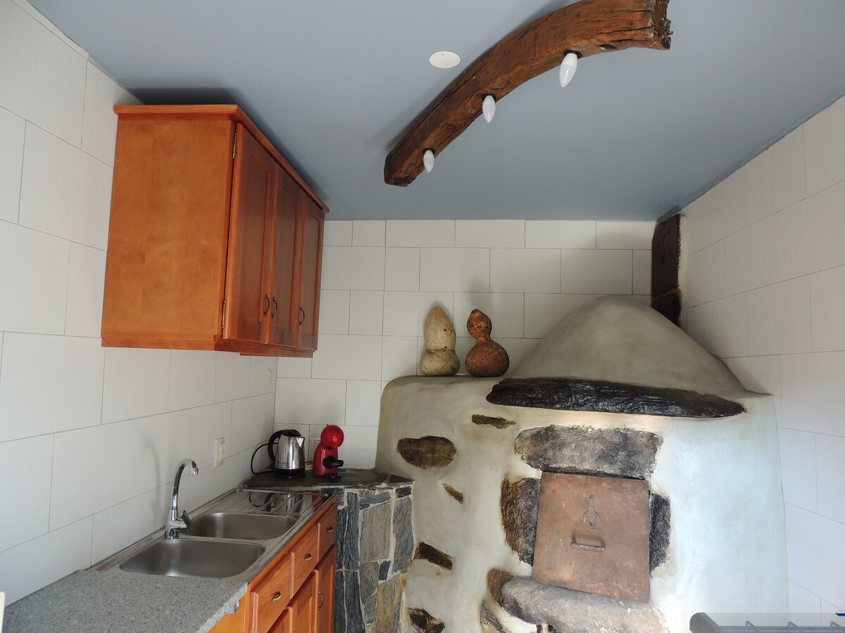 A functional kitchen is presented with wooden cabinets and a stone surface. An original baking oven, crafted from local materials, is featured prominently. Light fixtures are installed beneath a rustic wooden beam, adding to the warm ambiance of the space.