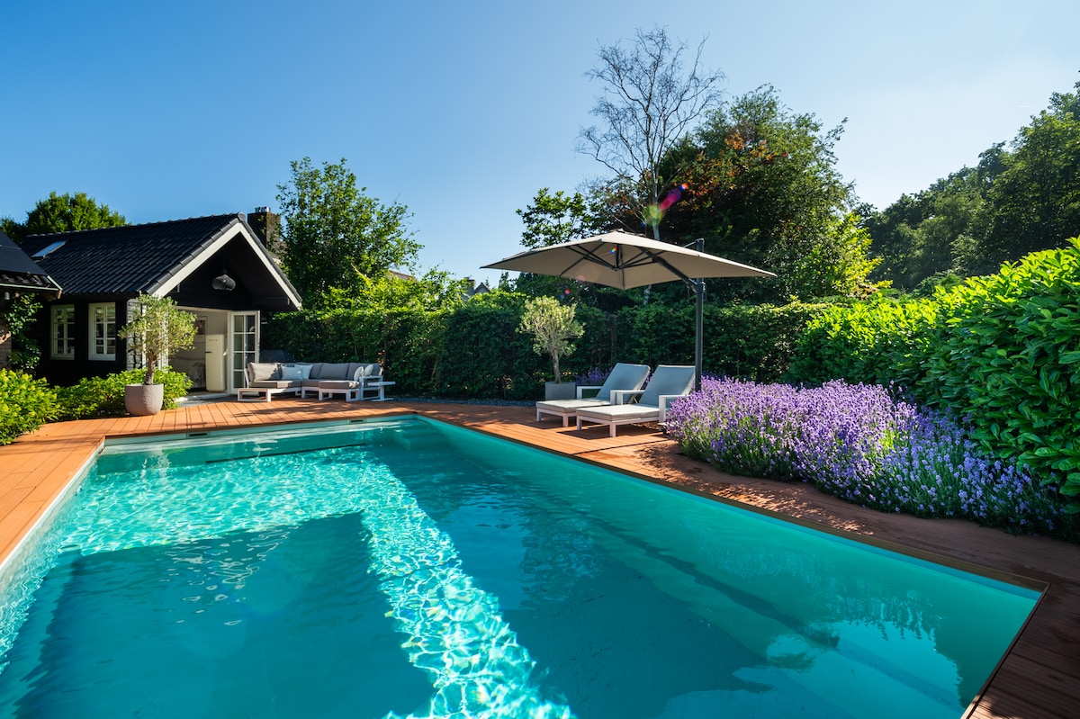 A tranquil pool area is highlighted, featuring a large swimming pool surrounded by a wooden deck. Two lounge chairs rest under a large umbrella, while vibrant lavender blooms add color to the lush greenery that encloses the space, providing privacy.