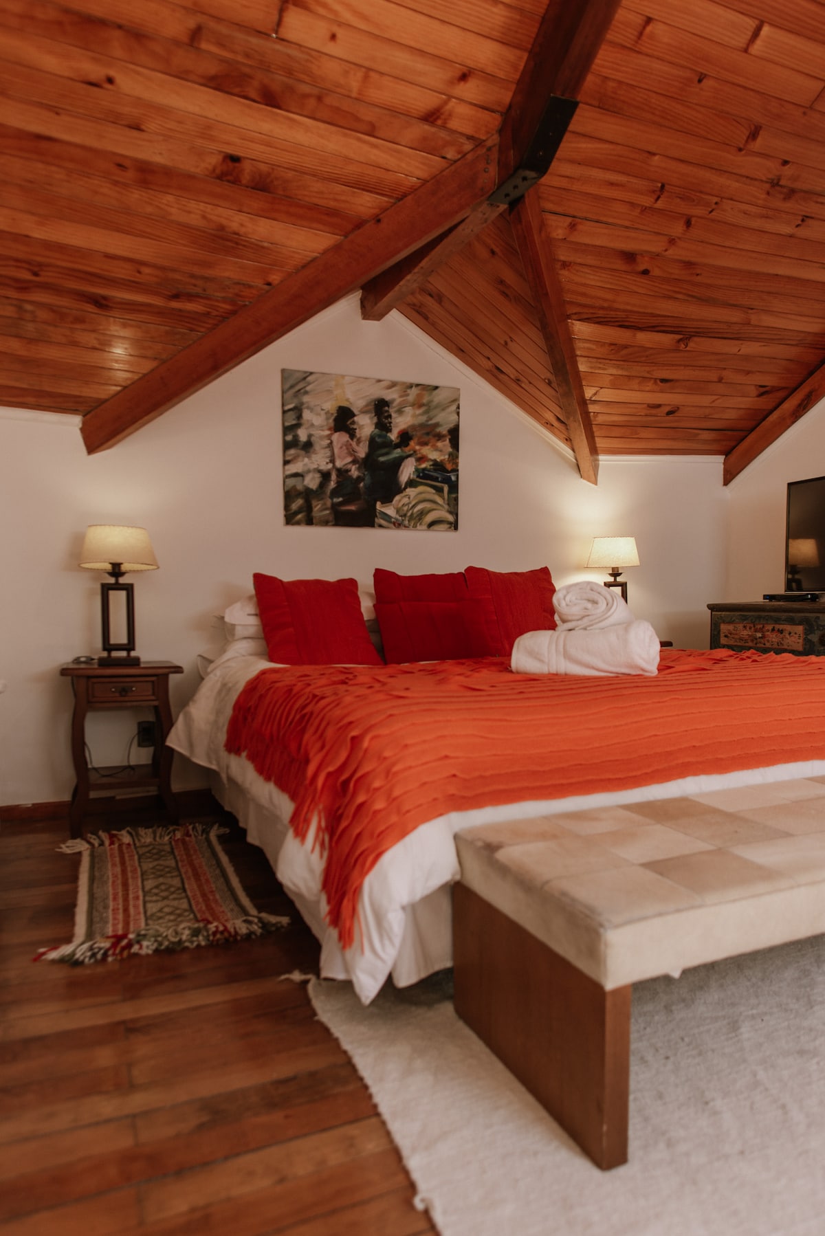 The room features a king-sized bed adorned with vibrant red pillows and a decorative towel sculpture. Warm wooden beams create a cozy atmosphere, complemented by bedside lamps and a stylish piece of art on the wall. A wooden bench and area rug enhance the inviting space.