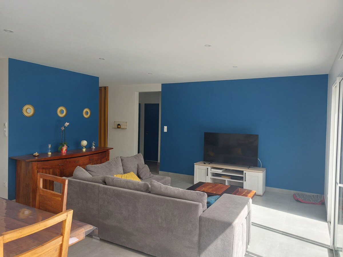 The living area is characterized by a modern design featuring a large gray sectional sofa and a coffee table. A TV is positioned on a media unit, with a blue accent wall creating a calming backdrop. Natural light flows through doorways leading to the exterior.
