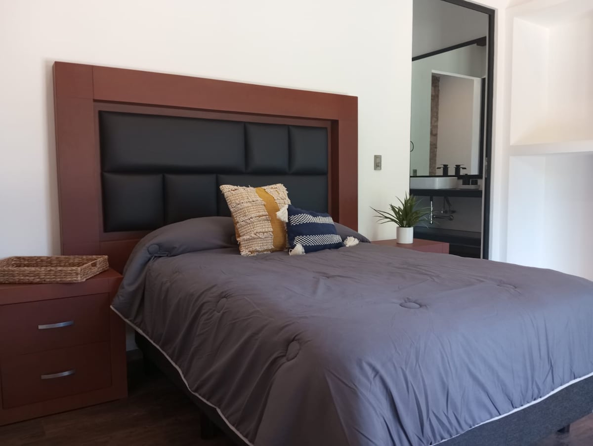 A spacious bed with a dark headboard is situated against a light-colored wall. Decorative pillows add subtle texture. A bedside table with drawers stands next to the bed, while a small potted plant introduces a hint of greenery. The bathroom area is visible in the background.