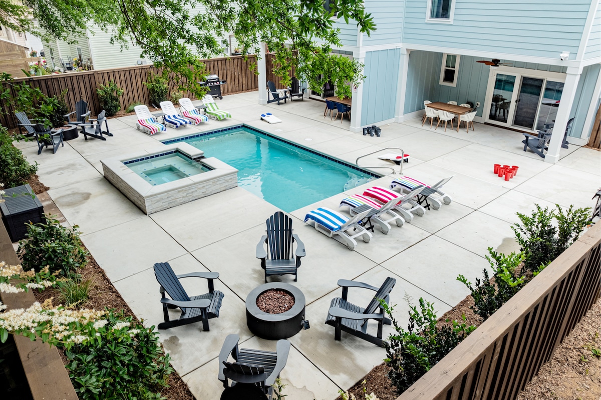 An inviting outdoor space features a large swimming pool surrounded by comfortable lounge chairs in various colors. A hot tub is situated nearby, along with a fire pit surrounded by adirondack chairs. Several outdoor dining areas are visible, creating a perfect setting for relaxation.