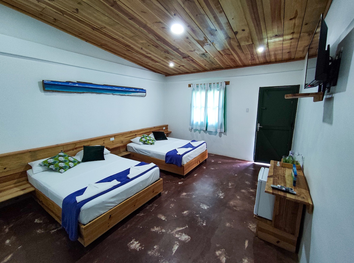 A spacious room features two wooden-framed beds, each adorned with white linens and blue accents. Natural light enters through a window, partially dressed with sheer curtains. A wooden table is situated against the wall, complemented by a television mounted above.