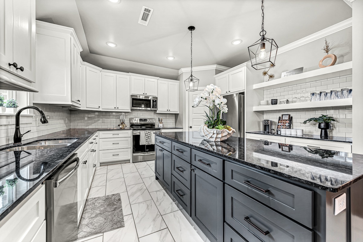 A modern kitchen is presented with white cabinetry and dark countertops. Stainless steel appliances are visible, alongside a large island that features a floral arrangement. Bright light floods the space, highlighting the tile flooring and the elegant design elements.