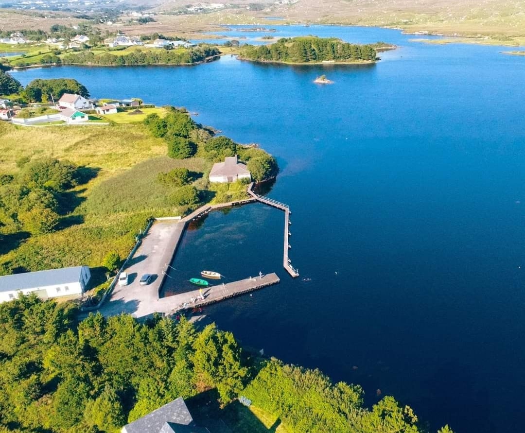 An aerial view showcases the calm waters of Loughanure lake, framed by lush greenery and rural landscapes. A private jetty extends into the water, with a few boats visible. The surrounding area includes patches of land and small clusters of buildings, all reflecting a peaceful environment.