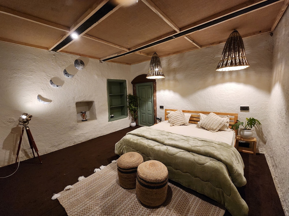 A spacious bedroom features a wooden bed adorned with a soft green blanket. Two decorative woven stools are positioned nearby. Soft lighting illuminates the room, complemented by unique pendant lamps. A small plant and minimalist decor enhance the serene atmosphere.